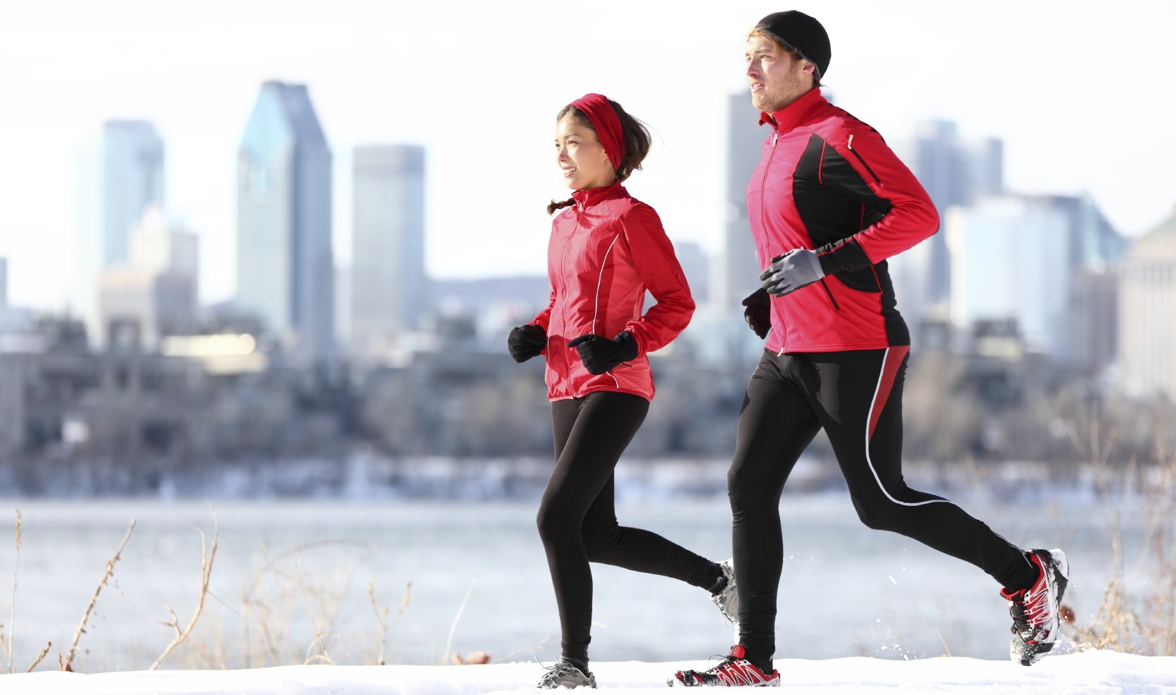 Runners running in winter snow with city skyline background. Healthy multiracial young couple. Asian woman runner and Caucasian man running with Montreal skyline, Quebec, Canada